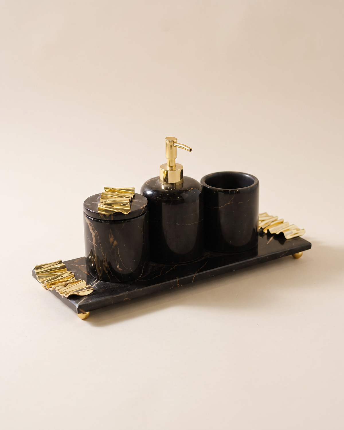 Four-piece black marble bath set with gold wave accents - tray, lidded box, soap dispenser and toothbrush holder - on a pale ivory studio background