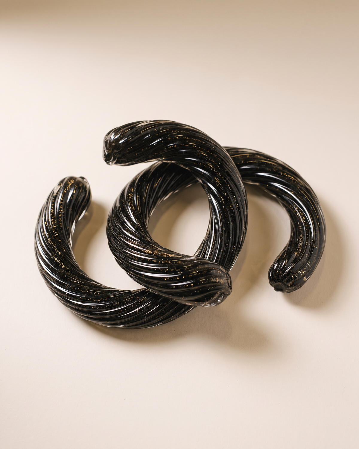 Two interlocking Murano glass chain links in black glass with gold flecks, pale ivory studio background